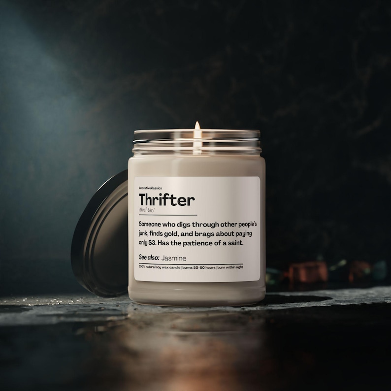 May include: A lit, white candle in a glass jar with a silver lid. The label reads "Thrifter" with a definition and "See also: Jasmine." The candle is on a dark surface.