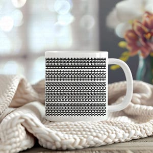 Pu&ograve; includere: Tazza in ceramica bianca con un motivo nero ripetuto di forme geometriche. La tazza ha un manico curvo ed &egrave; appoggiata su una coperta di maglia color crema. Lo sfondo &egrave; sfocato, con accenni di fiori.