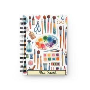 Puede incluir: Una libreta de espiral blanca con un patr&oacute;n colorido de pinceles, paletas de pintura y otros materiales de arte. La libreta tiene una etiqueta amarilla con el texto "Mrs. Smith" en ella.