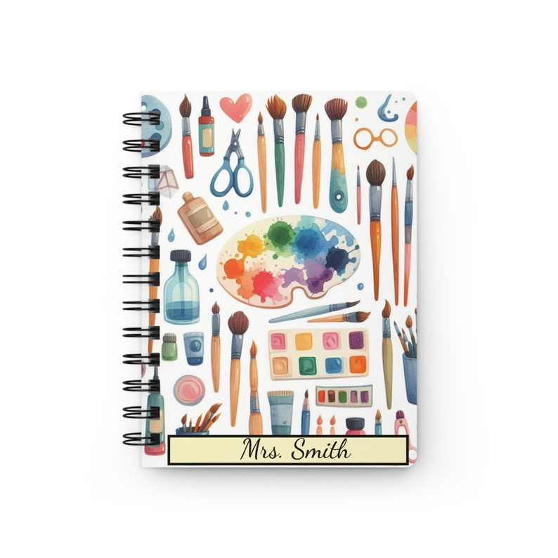 Puede incluir: Una libreta de espiral blanca con un patr&oacute;n colorido de pinceles, paletas de pintura y otros materiales de arte. La libreta tiene una etiqueta amarilla con el texto "Mrs. Smith" en ella.