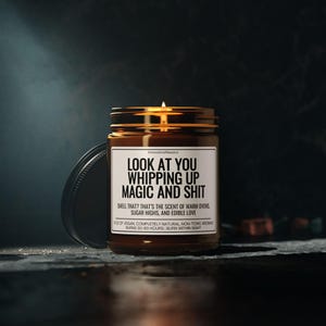 K&ouml;nnte beinhalten: Eine brennende, braune Glas-Kerze mit schwarzem Deckel. Auf dem Etikett steht "LOOK AT YOU WHIPPING UP MAGIC AND SHIT". Der Text darunter lautet: "SMELL THAT? THAT'S THE SCENT OF WARM OVENS, SUGAR HIGHS, AND EDIBLE LOVE." Die Kerze brennt 50-60 Stunden lang.