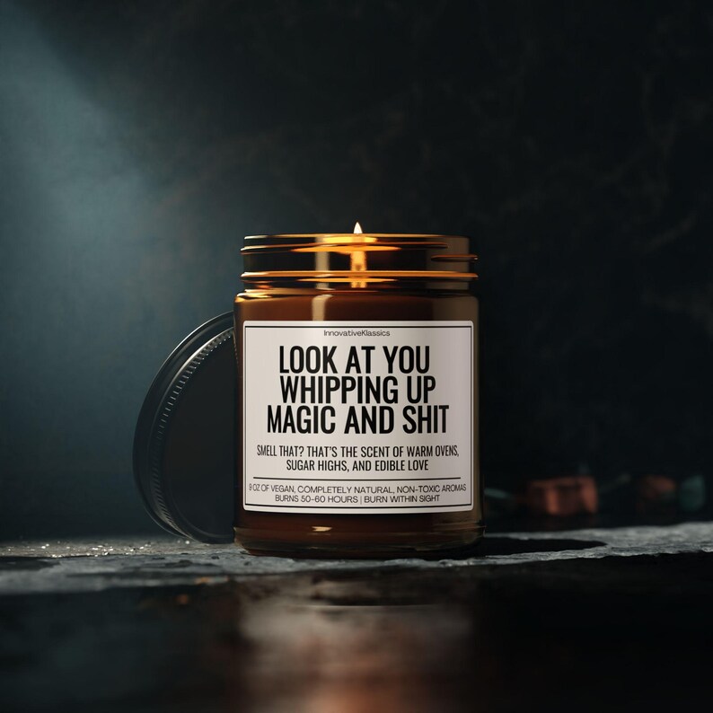 K&ouml;nnte beinhalten: Eine brennende, braune Glas-Kerze mit schwarzem Deckel. Auf dem Etikett steht "LOOK AT YOU WHIPPING UP MAGIC AND SHIT". Der Text darunter lautet: "SMELL THAT? THAT'S THE SCENT OF WARM OVENS, SUGAR HIGHS, AND EDIBLE LOVE." Die Kerze brennt 50-60 Stunden lang.