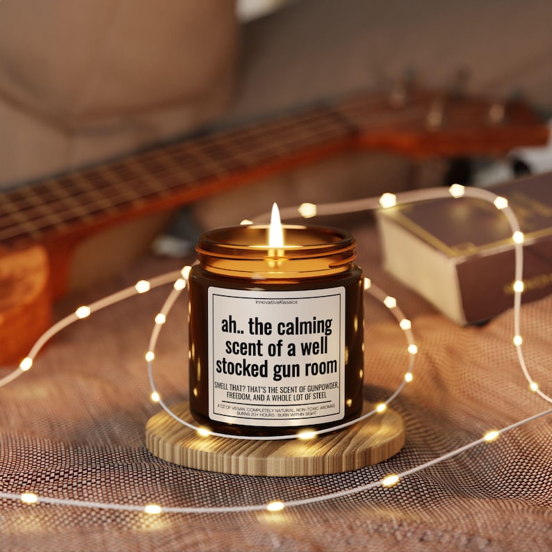 Puede incluir: Una vela de vidrio marr&oacute;n con una etiqueta blanca que dice "ah.. the calming scent of a well stocked gun room" y "SMELL THAT? THAT'S THE SCENT OF GUNPOWDER FREEDOM, AND A WHOLE LOT OF STEEL". La vela est&aacute; encendida y se encuentra sobre un posavasos de madera.