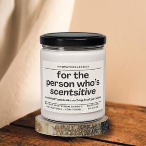 May include: A white glass jar candle with a black lid, resting on a wooden coaster. The label reads "for the person who's scentsitive." The candle is 10 oz soy wax, vegan, natural, and non-toxic, with a burn time of 50-60 hours.