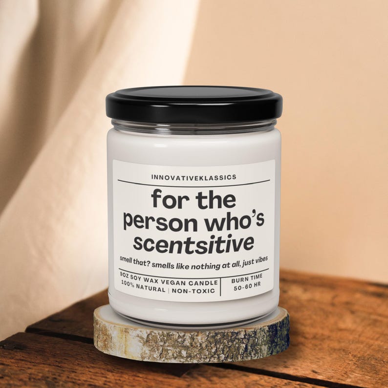 May include: A white glass jar candle with a black lid, resting on a wooden coaster. The label reads "for the person who's scentsitive." The candle is 10 oz soy wax, vegan, natural, and non-toxic, with a burn time of 50-60 hours.