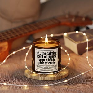 May include: A lit brown glass candle jar with a gold lid, sitting on a wooden coaster. The candle has a white label with black text that reads "ah.. the calming scent of ripping open a fresh pack of cards". Fairy lights and a ukulele are in the background.