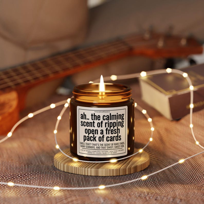May include: A lit brown glass candle jar with a gold lid, sitting on a wooden coaster. The candle has a white label with black text that reads "ah.. the calming scent of ripping open a fresh pack of cards". Fairy lights and a ukulele are in the background.