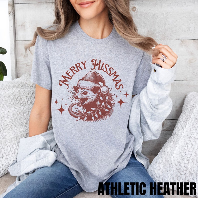 K&ouml;nnte beinhalten: Ein graues T-Shirt mit einer roten Grafik eines Opossums, das einen Weihnachtsmann-Hut tr&auml;gt und Weihnachtslichter h&auml;lt. Der Text "Merry Hisssmas" ist in roter Kursivschrift um das Opossum geschrieben.