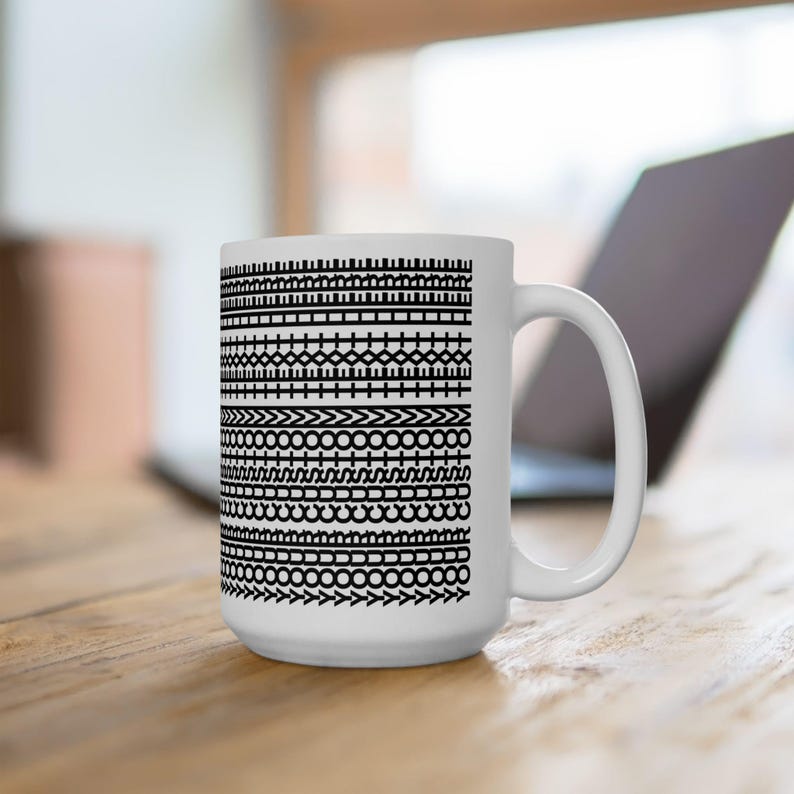 Pu&ograve; includere: Tazza in ceramica bianca con un motivo nero ripetuto di forme e linee geometriche. Il design include file di quadrati, cerchi e frecce. La tazza ha un manico grande ed &egrave; appoggiata su una superficie di legno.