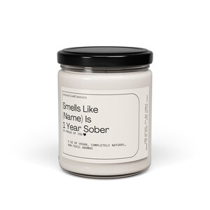 May include: A white candle jar with a black lid. The label reads "Smells Like (Name) Is 1 Year Sober" with a heart symbol. The label also says "9 oz of vegan, completely natural, non-toxic aromas".