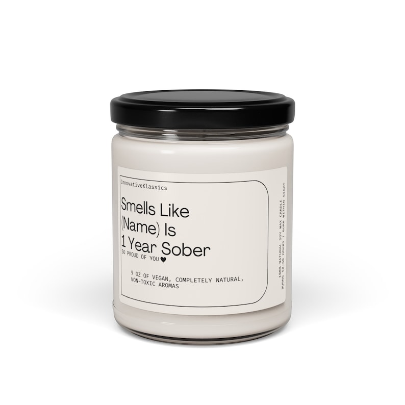 May include: A white candle jar with a black lid. The label reads "Smells Like (Name) Is 1 Year Sober" with a heart symbol. The label also says "9 oz of vegan, completely natural, non-toxic aromas".
