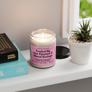 May include: A white candle with a pink label that reads "Fueled By Hot Flashes & Mood Swings" and "Who knew this would be my hot phase?" The candle is in a glass jar with a black lid. The candle is lit and there is a small plant in a white pot in the background.