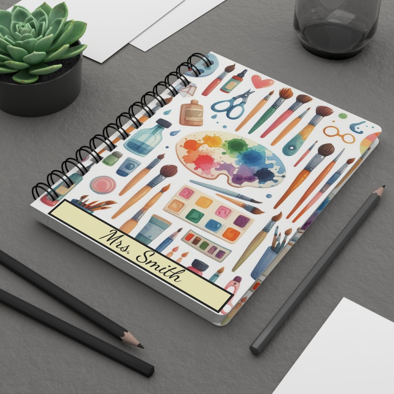Puede incluir: Una libreta de espiral blanca con un dise&ntilde;o colorido de pinceles, paletas de pintura y otros materiales de arte. La libreta tiene una etiqueta amarilla con el texto "Mrs. Smith" en ella.