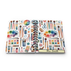 Puede incluir: Un cuaderno blanco de espiral con un patr&oacute;n colorido de art&iacute;culos de arte, incluyendo pinceles, paletas de pintura y tijeras. El cuaderno tiene una etiqueta amarilla en la parte delantera que dice "Sra. Smith".