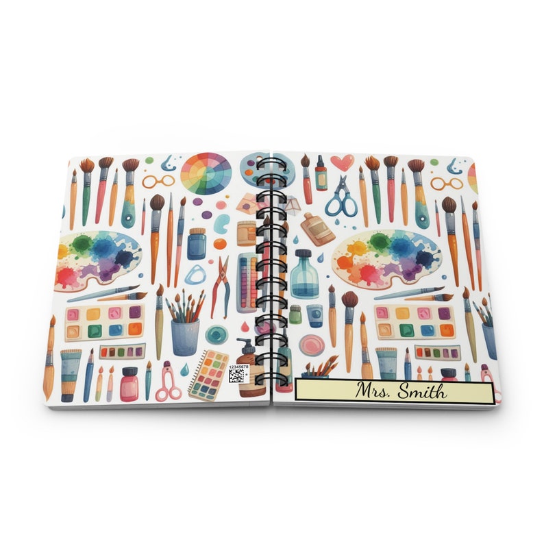 Puede incluir: Un cuaderno blanco de espiral con un patr&oacute;n colorido de art&iacute;culos de arte, incluyendo pinceles, paletas de pintura y tijeras. El cuaderno tiene una etiqueta amarilla en la parte delantera que dice "Sra. Smith".