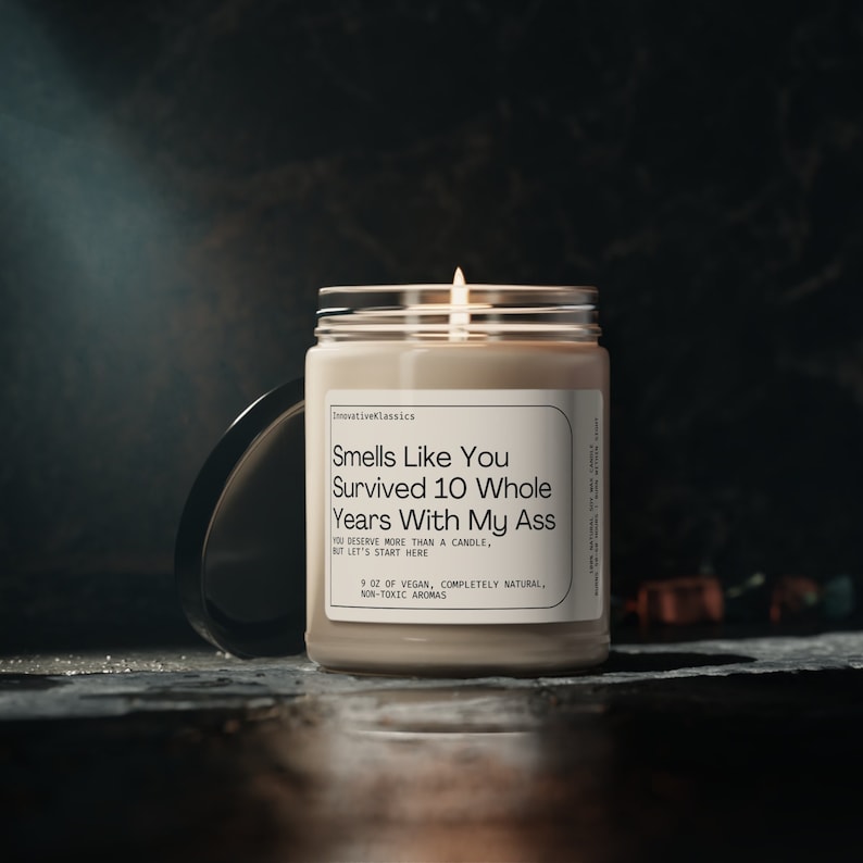 May include: A beige candle with a black label that reads "Smells Like You Survived 10 Whole Years With My Ass". The label also includes the text "You deserve more than a candle, but let's start here. 9 oz of vegan, completely natural, non-toxic aromas."