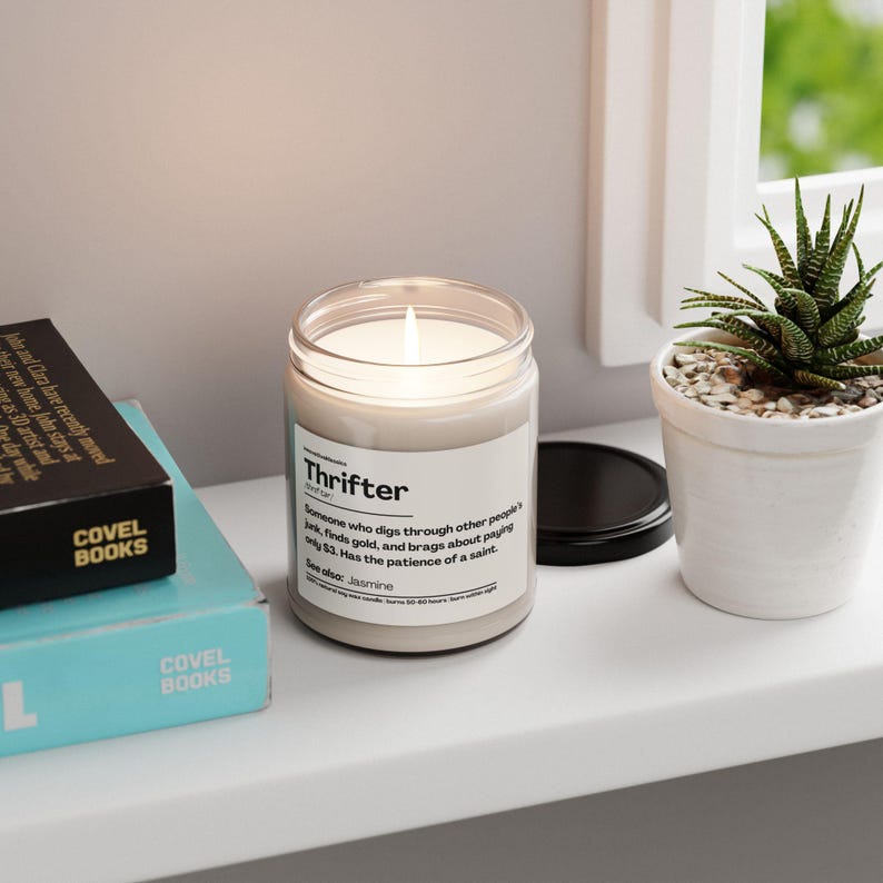 May include: A lit, scented candle in a clear glass jar with a black lid. The label reads "Thrifter" with a definition. A small potted succulent and two books are also visible.