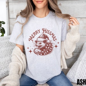 K&ouml;nnte beinhalten: Ein graues T-Shirt mit einer roten Grafik eines Opossums, das einen Weihnachtsmann-Hut tr&auml;gt und von Weihnachtslichtern umgeben ist. Der Text "Merry Hissmas" ist in einem roten Kreis um das Opossum geschrieben.