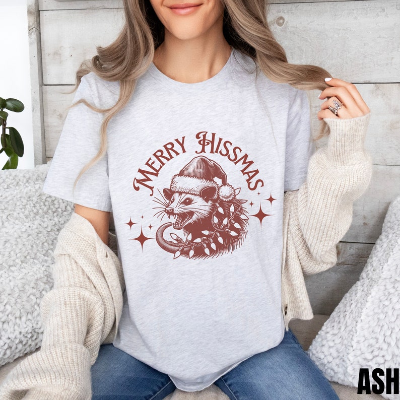 K&ouml;nnte beinhalten: Ein graues T-Shirt mit einer roten Grafik eines Opossums, das einen Weihnachtsmann-Hut tr&auml;gt und von Weihnachtslichtern umgeben ist. Der Text "Merry Hissmas" ist in einem roten Kreis um das Opossum geschrieben.