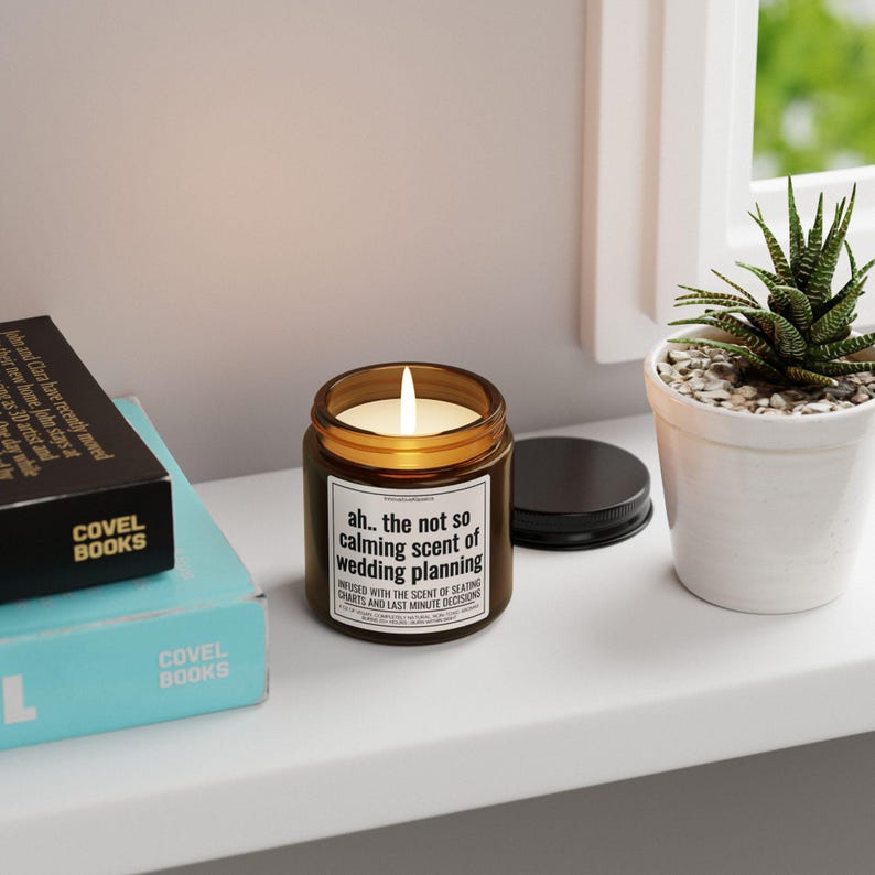 May include: A brown glass candle with a white label that reads "ah.. the not so calming scent of wedding planning. Infused with the scent of seating charts and last minute decisions."