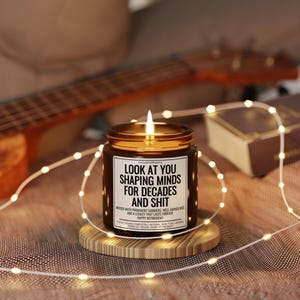 Pode incluir: Uma vela de vidro castanho acesa com tampa dourada, sobre um descanso de madeira. A vela tem um r&oacute;tulo branco com texto preto: "LOOK AT YOU SHAPING MINDS FOR DECADES AND SHIT". Luzes de fadas e um ukulele no fundo.