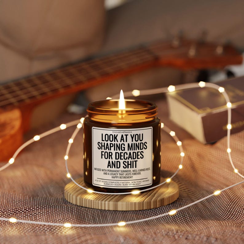 Pode incluir: Uma vela de vidro castanho acesa com tampa dourada, sobre um descanso de madeira. A vela tem um r&oacute;tulo branco com texto preto: "LOOK AT YOU SHAPING MINDS FOR DECADES AND SHIT". Luzes de fadas e um ukulele no fundo.