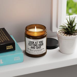 Puede incluir: Una vela encendida en un tarro de vidrio marr&oacute;n con una etiqueta blanca que dice "LOOK AT YOU CUTTING HAIR AND SHIT". La vela est&aacute; en un estante blanco junto a una peque&ntilde;a suculenta en maceta y dos libros.