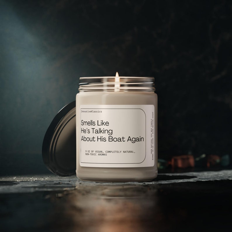 Op de afbeelding: Een beige kaars in een glazen pot met een wit etiket waarop staat "Smells Like He's Talking About His Boat Again". Het etiket vermeldt ook "9 oz Vegan, volledig natuurlijk, niet-giftige aroma's".