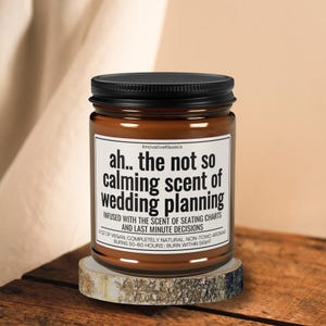 May include: A brown glass candle jar with a black lid. The label reads "ah.. the not so calming scent of wedding planning" and "INFUSED WITH THE SCENT OF SEATING CHARTS AND LAST MINUTE DECISIONS". The candle is sitting on a wooden surface.