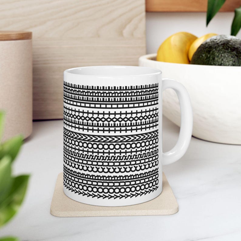 Pu&ograve; includere: Tazza in ceramica bianca con un motivo geometrico nero. Il design presenta bande orizzontali di varie forme, tra cui linee, croci e cerchi. La tazza &egrave; appoggiata su un sottobicchiere beige.