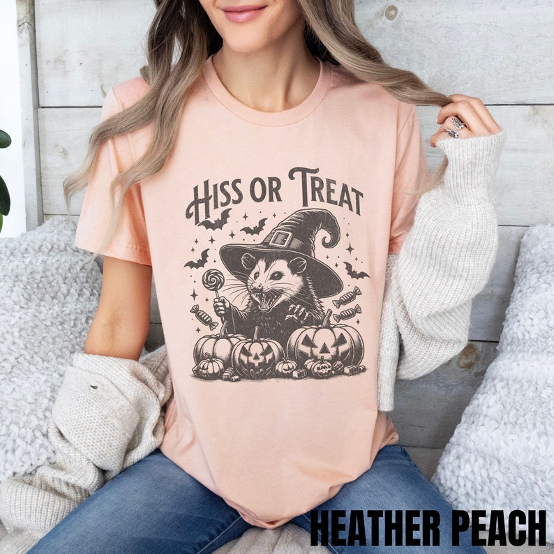May include: A peach-colored t-shirt with a black and white illustration of a possum wearing a witch's hat and holding a lollipop. The text "Hiss or Treat" is above the possum. The possum is surrounded by candy and pumpkins.