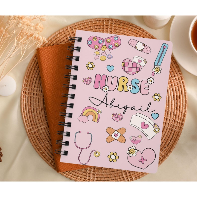 Op de afbeelding: Roze spiraalnotitieblok met een schattig cartoonontwerp met een stethoscoop, verband, harten en bloemen. De tekst "NURSE Abigail" staat op de omslag.