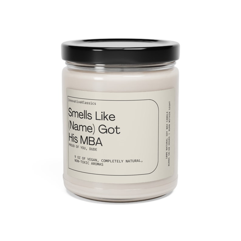 K&ouml;nnte beinhalten: Eine Glaskerze in einem Glas mit schwarzem Deckel. Das Etikett lautet "Smells Like (Name) Got His MBA. Proud of You, Dude. 9 oz of Vegan, Completely Natural, Non-Toxic Aromas."
