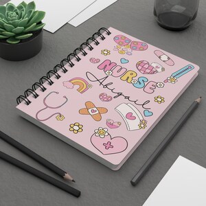 Op de afbeelding: Roze spiraalnotitieblok met een kleurrijk ontwerp met het woord "VERPLEEGKUNDIGE" en de naam "Abigail". Het ontwerp bevat harten, sterren, regenbogen, verbanden, een stethoscoop en een verpleegstersmuts.