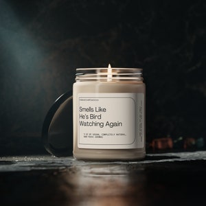 May include: A beige candle in a glass jar with a white label that reads "Smells Like He's Bird Watching Again". The label also says "9 oz of vegan, completely natural, non-toxic aromas".