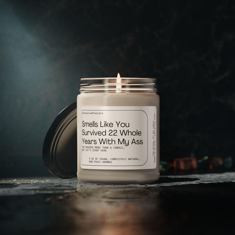 May include: A beige candle with a black lid. The candle has a white label with the text "Smells Like You Survived 22 Whole Years With My Ass" and "5 oz of vegan, completely natural, non-toxic aromas".