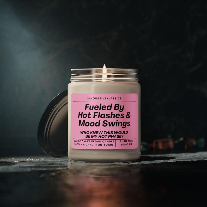 May include: A white candle with a pink label that reads "Fueled By Hot Flashes & Mood Swings" and "Who knew this would be my hot phase?" The candle is a soy wax vegan candle and is 100% natural and non-toxic. The candle has a burn time of 50-60 hours.