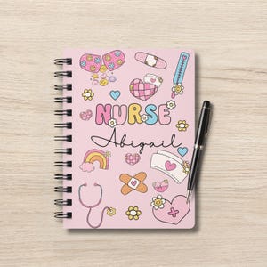 Op de afbeelding: Een roze notitieboek met spiraalbinding en een kleurrijk cartoonontwerp met een stethoscoop, verband, harten en de woorden "NURSE Abigail".