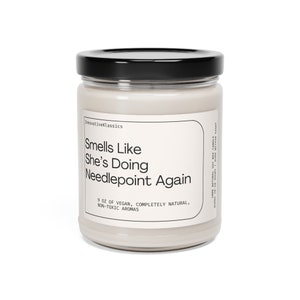 May include: A glass jar candle with a black lid and a white label that reads "Smells Like She's Doing Needlepoint Again". The label also says "9 oz of vegan, completely natural, non-toxic aromas".