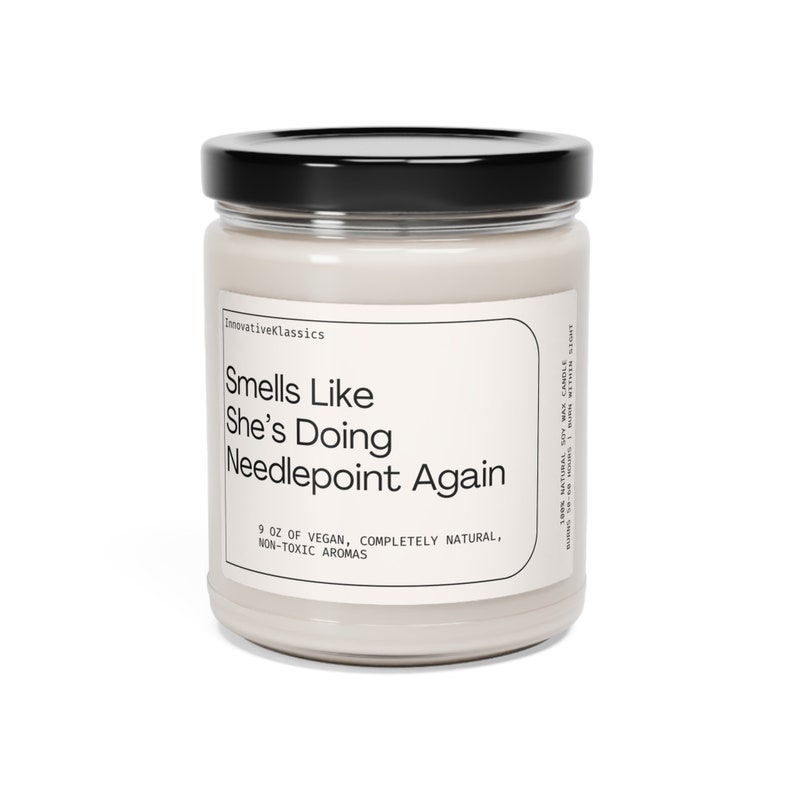 May include: A glass jar candle with a black lid and a white label that reads "Smells Like She's Doing Needlepoint Again". The label also says "9 oz of vegan, completely natural, non-toxic aromas".