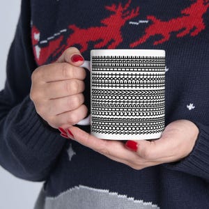 Pu&ograve; includere: Una tazza di ceramica bianca con un motivo geometrico nero. Il motivo &egrave; composto da varie bande orizzontali di linee, punti e frecce. La tazza &egrave; tenuta da una persona che indossa un maglione blu navy.