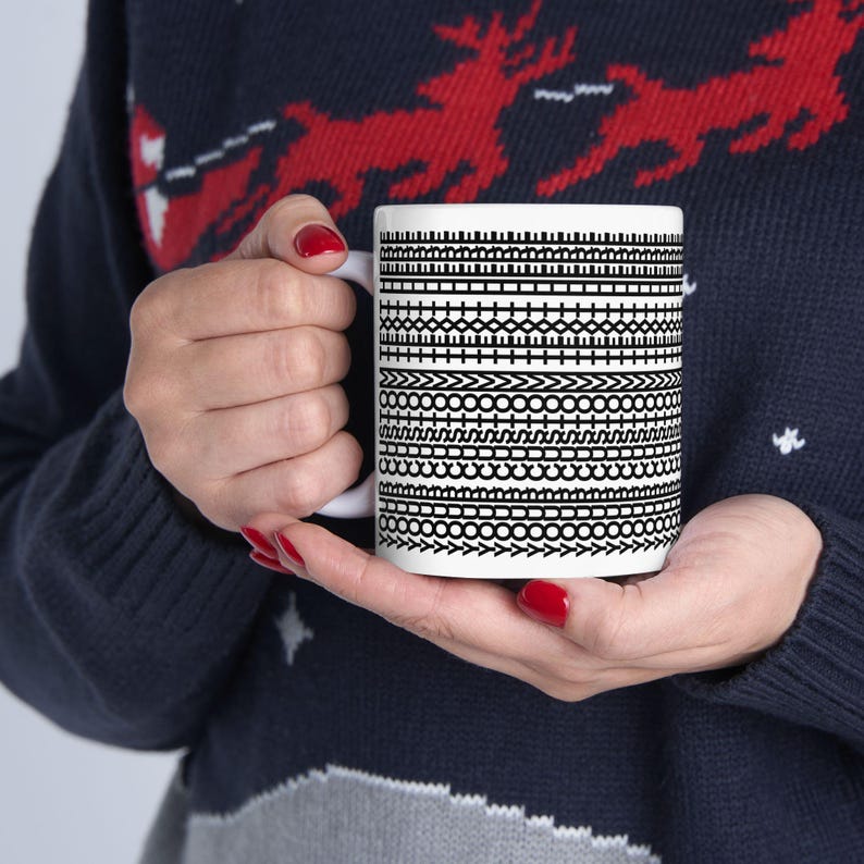 Pu&ograve; includere: Una tazza di ceramica bianca con un motivo geometrico nero. Il motivo &egrave; composto da varie bande orizzontali di linee, punti e frecce. La tazza &egrave; tenuta da una persona che indossa un maglione blu navy.