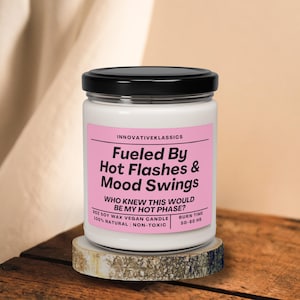 May include: A white candle with a black lid and a pink label that reads "Fueled By Hot Flashes & Mood Swings. Who knew this would be my hot phase?" 8 oz soy wax vegan candle. 100% natural | non-toxic. Burn time 50-60 hr.