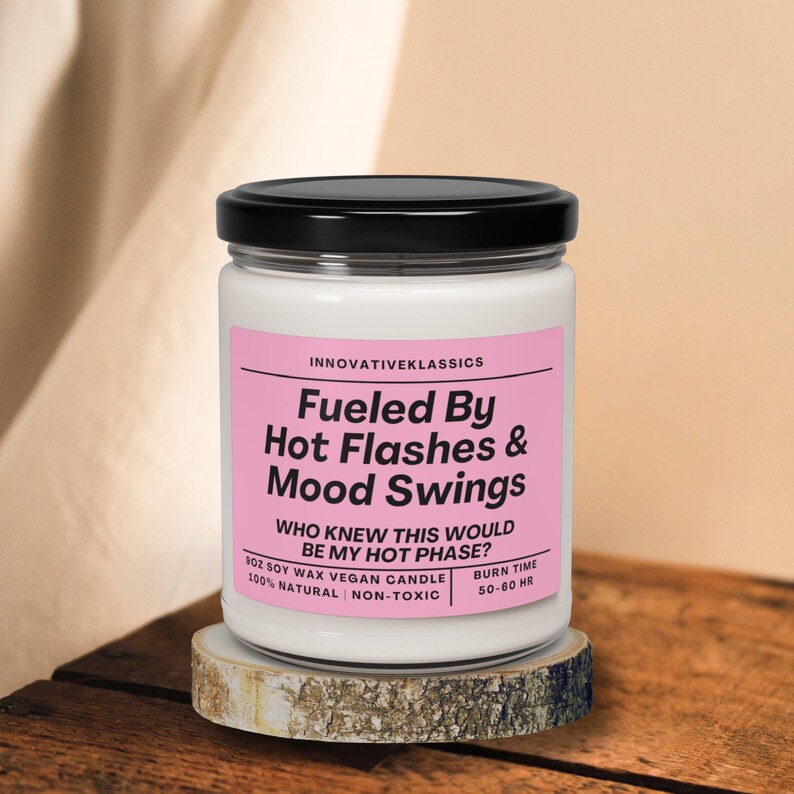 May include: A white candle with a black lid and a pink label that reads "Fueled By Hot Flashes & Mood Swings. Who knew this would be my hot phase?" 8 oz soy wax vegan candle. 100% natural | non-toxic. Burn time 50-60 hr.