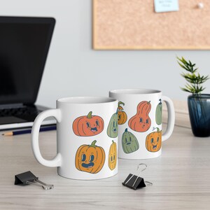 Op de afbeelding: Twee witte keramische mokken met een Halloween-thema. De mokken hebben verschillende cartoon-pompoenen met verschillende gezichtsuitdrukkingen en kleuren. De mokken staan op een lichtbruin houten oppervlak.