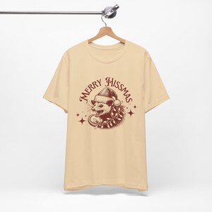 K&ouml;nnte beinhalten: Ein beiges T-Shirt mit einer roten Grafik eines Opossums, das einen Weihnachtsmann-Hut und eine Girlande tr&auml;gt. Der Text "Merry Hissmas" ist in roter Kursivschrift um das Opossum geschrieben.
