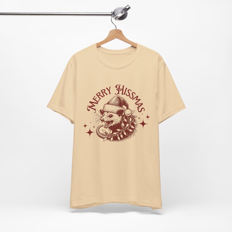 K&ouml;nnte beinhalten: Ein beiges T-Shirt mit einer roten Grafik eines Opossums, das einen Weihnachtsmann-Hut und eine Girlande tr&auml;gt. Der Text "Merry Hissmas" ist in roter Kursivschrift um das Opossum geschrieben.