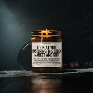 May include: A lit candle in a brown glass jar with a black lid. The label reads "LOOK AT YOU MASTERING THE STOCK MARKET AND SHIT." The text below says "SMELL THAT? THAT'S THE SCENT OF LATE-NIGHT TRADES, MARKET SWINGS, AND MILD REGRET."