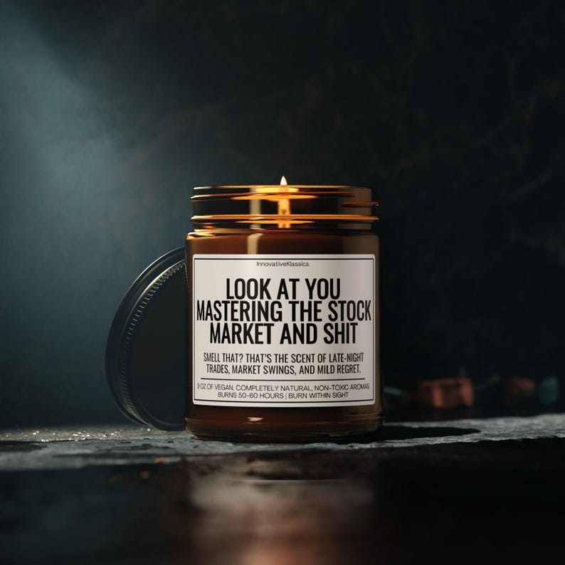 May include: A lit candle in a brown glass jar with a black lid. The label reads "LOOK AT YOU MASTERING THE STOCK MARKET AND SHIT." The text below says "SMELL THAT? THAT'S THE SCENT OF LATE-NIGHT TRADES, MARKET SWINGS, AND MILD REGRET."