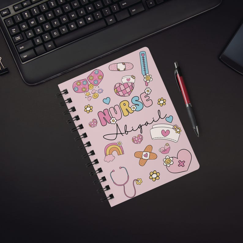 Op de afbeelding: Roze spiraalnotitieblok met een kleurrijk ontwerp met het woord "NURSE" en de naam "Abigail". Het notitieblok is versierd met harten, bloemen, verbanden, een stethoscoop en andere medische illustraties.